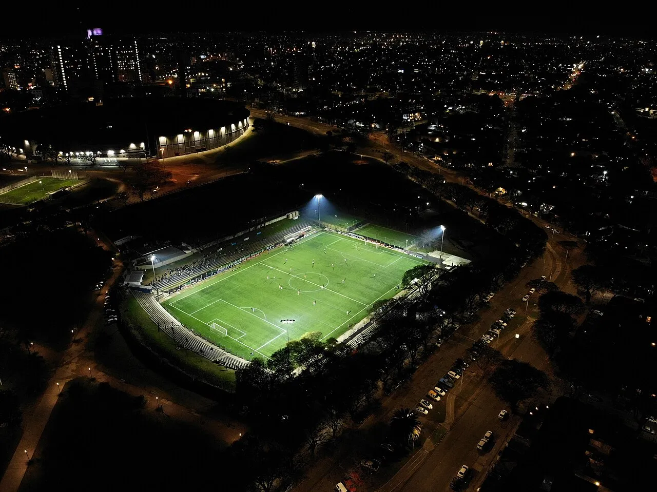 Vista aérea nocturna de un complejo deportivo con cancha de fútbol iluminada y un edificio circular de fondo, dentro del Parque Batlle, Montevideo