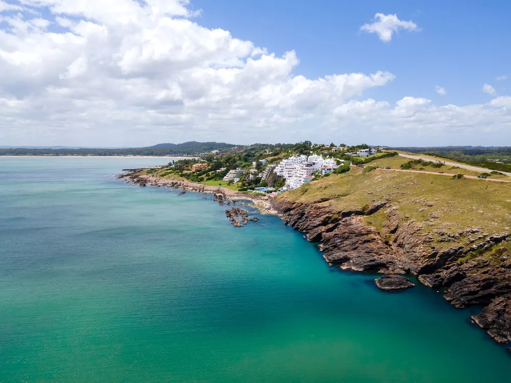 Vista aérea de la costa de Punta Ballena, con Casapueblo asomada sobre las rocas y el mar turquesa.
