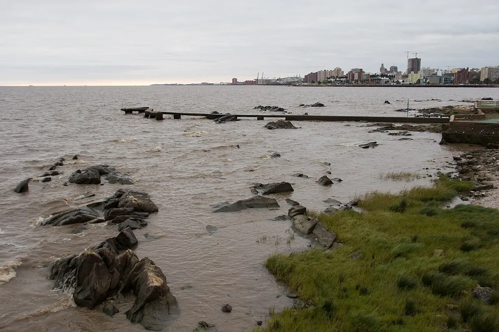 Costa rocosa de Punta Carretas con un antiguo espigón de madera deteriorado por el oleaje y el perfil de edificios de la costa al fondo